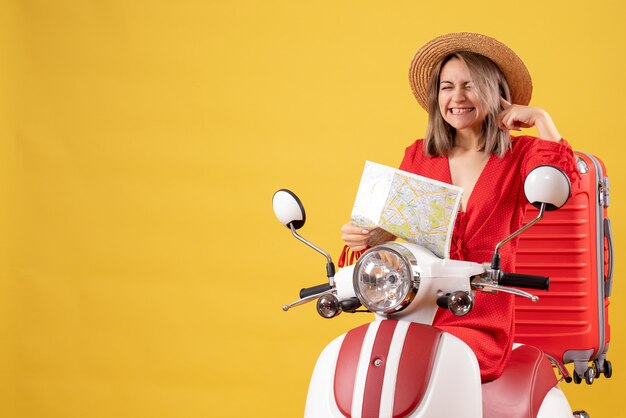 Chica guapa en ciclomotor con maleta roja sosteniendo mapa cerrando su oreja