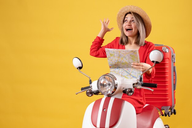 Chica guapa en ciclomotor con maleta roja sosteniendo mapa mirando hacia arriba