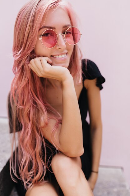 Chica guapa complacida con gafas de sol tocando su rostro y sonriendo.