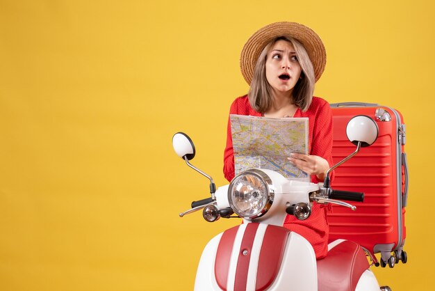 Chica guapa desconcertada en ciclomotor con maleta roja sosteniendo el mapa