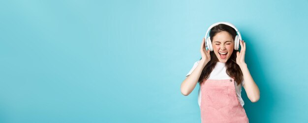 Chica guapa despreocupada grita de alegría escuchando música en auriculares con letras de sincronización de labios de cara feliz de pie sobre fondo azul