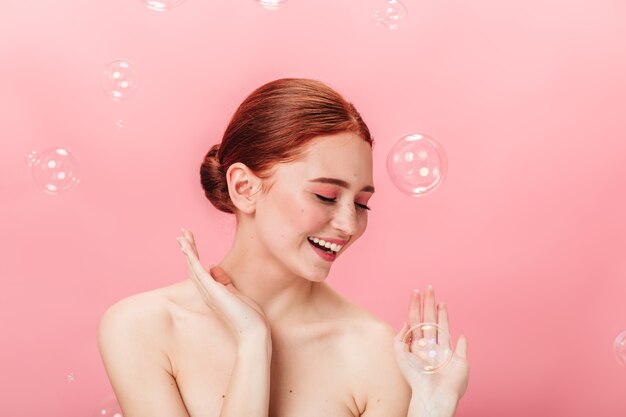 Chica guapa despreocupada posando con pompas de jabón. Disparo de estudio de atractiva mujer jengibre sonriendo sobre fondo rosa.