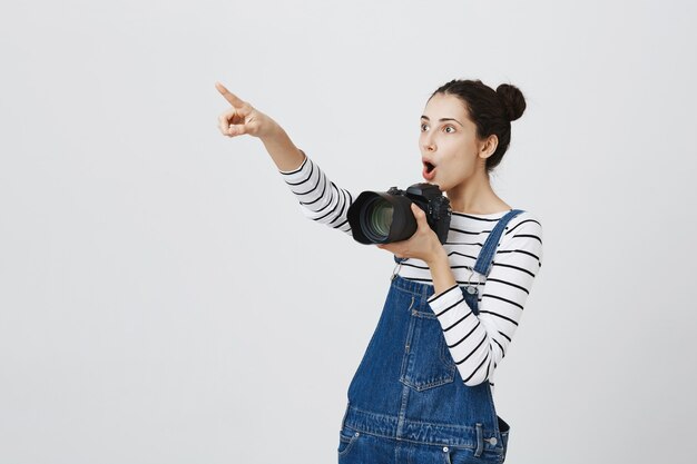 Chica guapa divertida apuntando hacia la esquina superior izquierda asombrada y sosteniendo la cámara, tomando fotografías