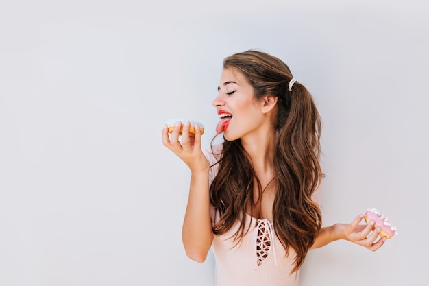 Chica guapa divertida con el pelo largo con donuts coloridos. Atractiva mujer joven en traje rosa lamiendo delicioso. Dulces, diversión, vida alegre.