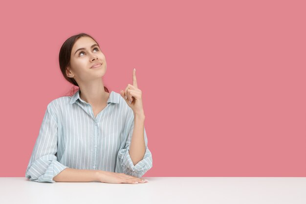 Foto gratuita chica guapa en el escritorio apuntando su dedo índice hacia arriba indicando una pared rosa en blanco para su texto o contenido publicitario. hembra joven alegre haciendo gesto con el dedo índice, mirando hacia arriba