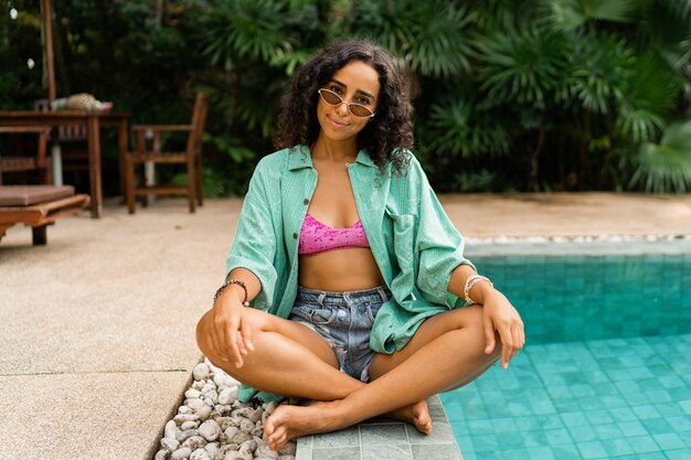 Chica guapa con estilo en traje de verano sentado cerca de la piscina Estado de ánimo de vacaciones y viajes Colores brillantes Accesorios de moda