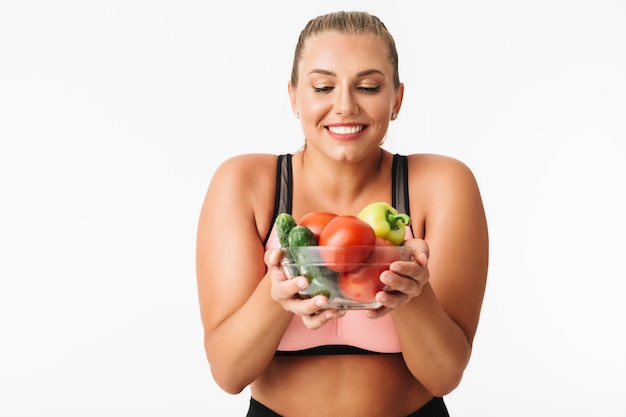 Chica guapa con exceso de peso en la parte superior deportiva felizmente sosteniendo un tazón con verduras en las manos sobre fondo blanco aislado