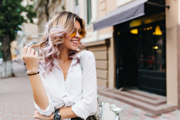 Chica guapa con gafas de sol y pulseras jugando con su pelo corto y rizado y sonriendo en la calle