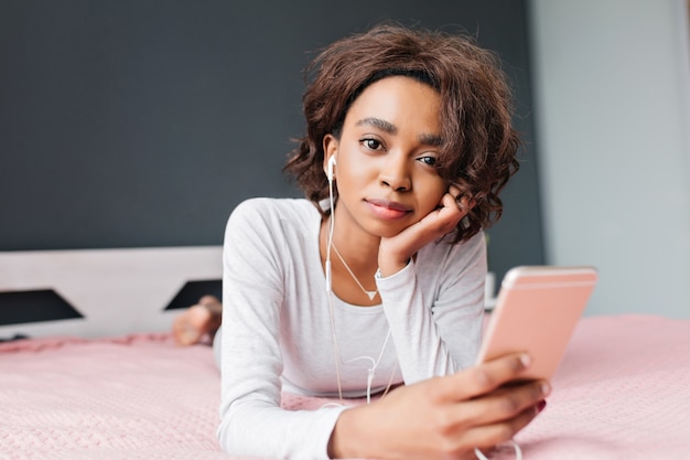 Chica guapa joven acostada en la cama, escuchando música en auriculares desde el teléfono inteligente rosa, descansando en casa. Vistiendo camiseta gris claro con mangas largas.