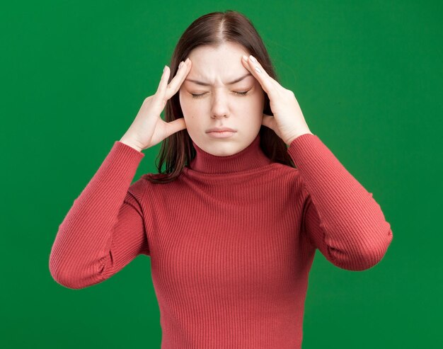 Chica guapa joven adolorida manteniendo las manos en la cabeza con los ojos cerrados aislados en la pared verde