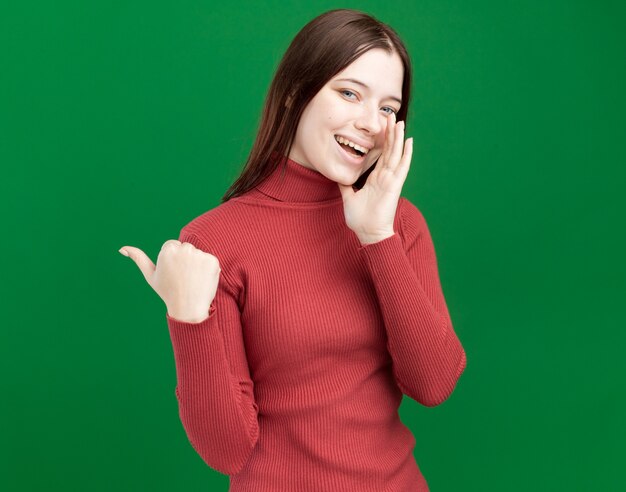 Chica guapa joven alegre apuntando al lado poniendo la mano cerca de la boca susurrando aislado en la pared verde con espacio de copia