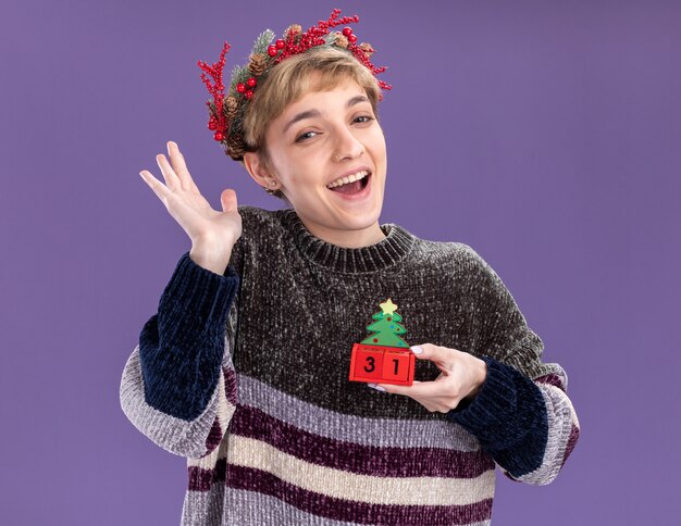 Chica guapa joven alegre con corona de cabeza de navidad sosteniendo el juguete del árbol de navidad con fecha mostrando la mano vacía aislada en la pared púrpura