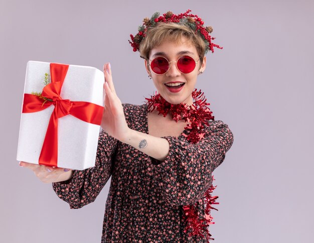 Chica guapa joven alegre con corona de Navidad y guirnalda de oropel alrededor del cuello con gafas estirando el paquete de regalo hacia aislado en la pared blanca con espacio de copia