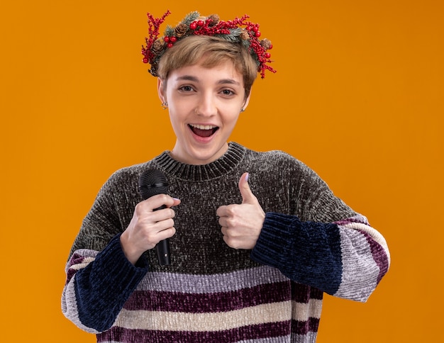 Chica guapa joven alegre con corona de Navidad sosteniendo el micrófono mostrando el pulgar hacia arriba aislado en la pared naranja