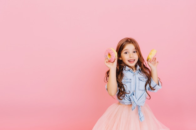 Chica guapa joven alegre emocionada en falda de tul expresando positividad, divirtiéndose a la cámara con donas aisladas sobre fondo rosa. Infancia feliz con delicioso postre. Colocar texto fot