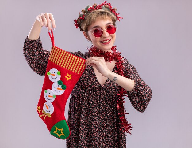 Chica guapa joven alegre con guirnalda de cabeza de navidad y guirnalda de oropel alrededor del cuello con gafas sosteniendo calcetín de navidad aislado en la pared blanca