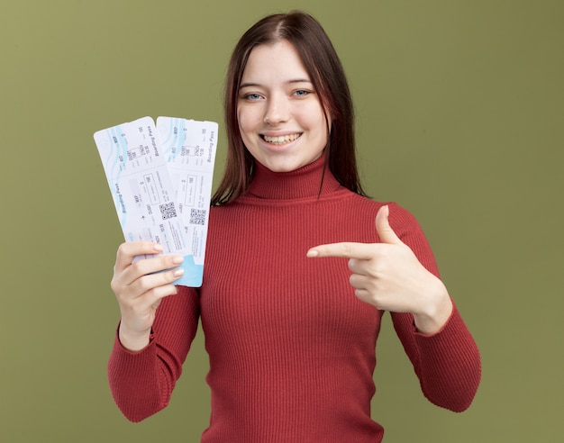 Chica guapa joven alegre sosteniendo y apuntando a los billetes de avión