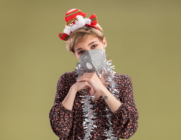 Chica guapa joven astuta con diadema de santa claus y guirnalda de oropel alrededor del cuello con máscara protectora mirando a la cámara manteniendo las manos juntas aisladas sobre fondo verde oliva