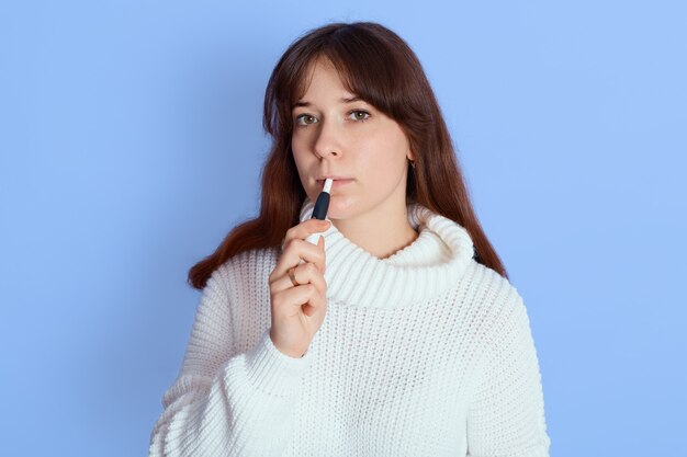 Chica guapa joven con cigarrillo electrónico posando sobre azul, mira a cámara, vistiendo un suéter blanco, cigarrillo electrónico de fumar mujer de pelo oscuro interior.