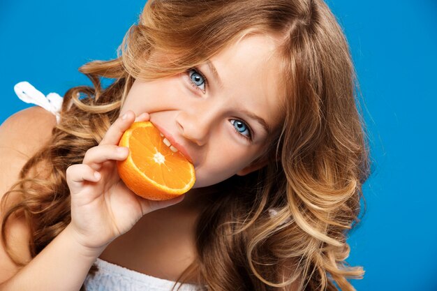 Chica guapa joven comiendo naranja sobre pared azul