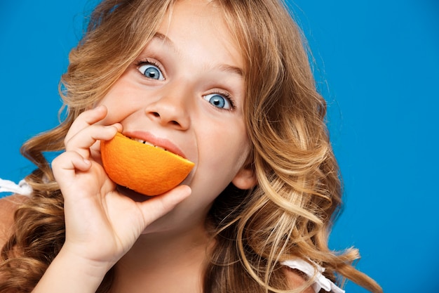 Chica guapa joven comiendo naranja sobre pared azul