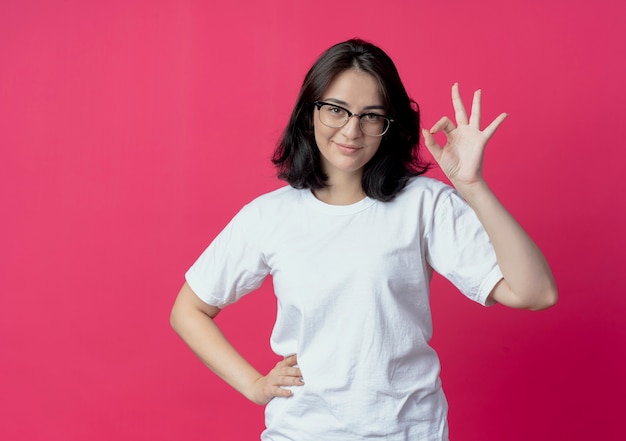 Chica guapa joven complacida con gafas poniendo la mano en la cintura y haciendo el signo de ok