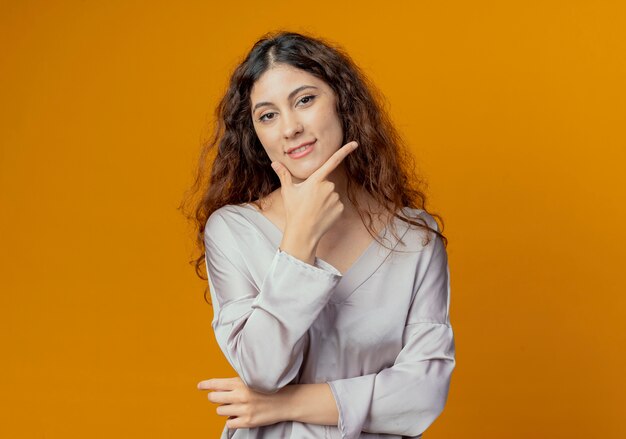 Chica guapa joven complacida poniendo la mano debajo de la barbilla aislada en la pared amarilla