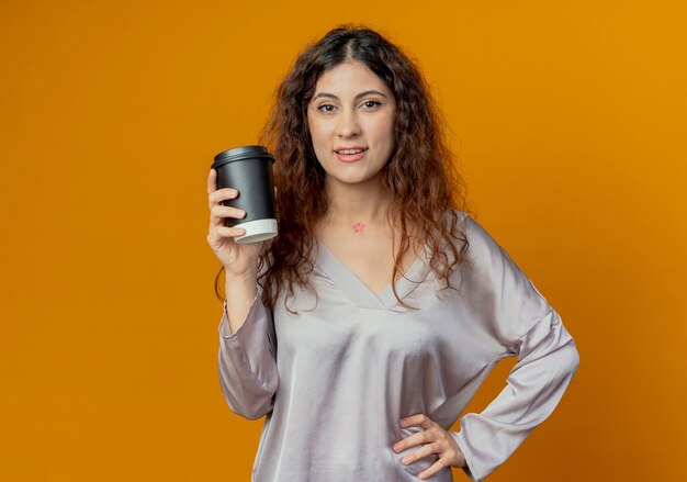 Chica guapa joven complacida sosteniendo una taza de café y poniendo la mano en la cadera aislada en la pared amarilla