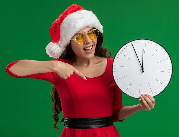 Chica guapa joven confiada con gorro de Papá Noel y gafas sosteniendo y apuntando al reloj mirando a cámara aislada sobre fondo verde