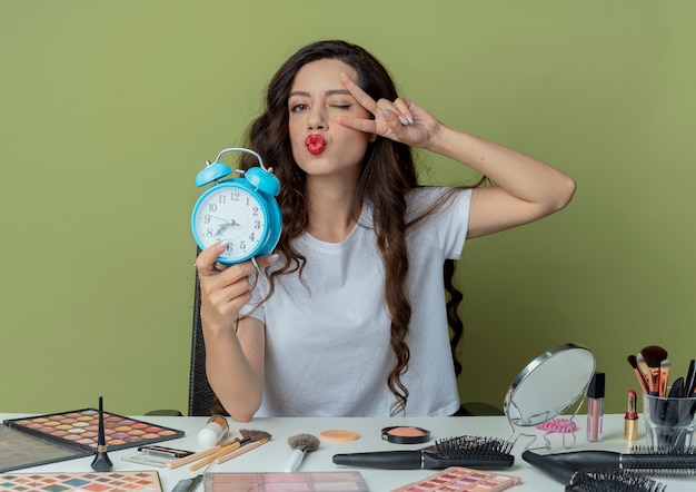 Chica guapa joven confiada que se sienta en la mesa de maquillaje con herramientas de maquillaje sosteniendo el despertador guiñando un ojo y haciendo el signo de la paz y mirando a la cámara a través de los dedos aislados sobre fondo verde oliva