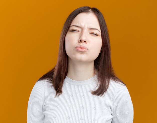 Chica guapa joven confundida entrecerrar los ojos con los labios fruncidos aislados en la pared naranja