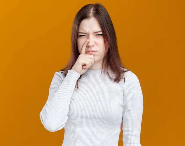 Chica guapa joven descontenta poniendo el dedo en la nariz aislado en la pared naranja