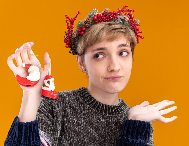 Chica guapa joven despistada con corona de cabeza de navidad sosteniendo adornos de navidad de santa claus mirando al lado mostrando la mano vacía aislada en la pared naranja