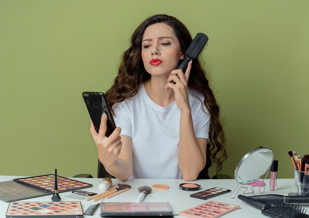 Chica guapa joven disgustada sentada en la mesa de maquillaje con herramientas de maquillaje sosteniendo y mirando el teléfono móvil y manteniendo el peine cerca de la oreja aislado sobre fondo verde oliva