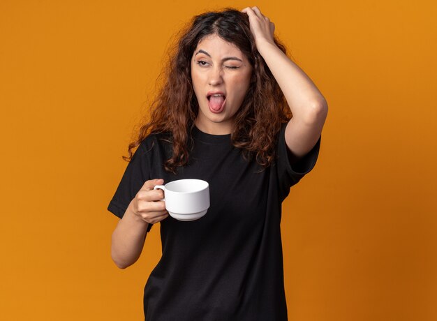 Chica guapa joven disgustada sosteniendo una taza de té manteniendo la mano en la cabeza mirando al lado mostrando la lengua con un ojo cerrado