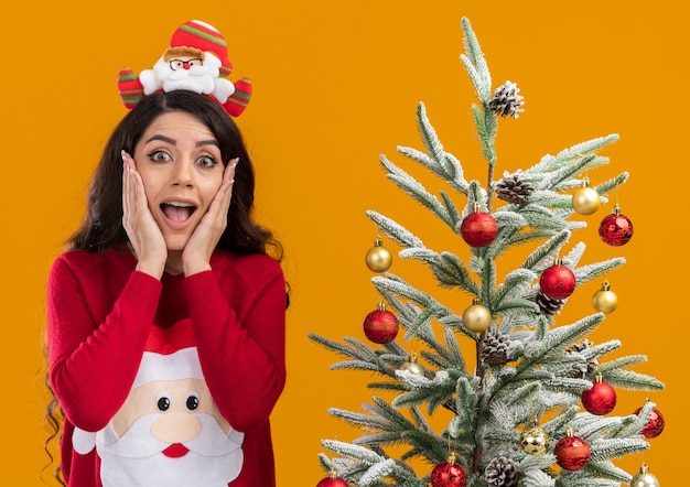 Chica guapa joven emocionada con diadema de santa claus y suéter de pie cerca del árbol de Navidad decorado manteniendo las manos en la cara mirando a cámara aislada sobre fondo naranja