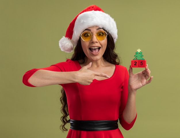 Chica guapa joven emocionada con gorro de Papá Noel y gafas sosteniendo y apuntando al juguete del árbol de Navidad con fecha mirando