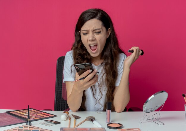 Chica guapa joven enojada sentada en la mesa de maquillaje con herramientas de maquillaje sosteniendo y mirando el teléfono móvil y gritando con pincel para polvos en otra mano aislada sobre fondo carmesí