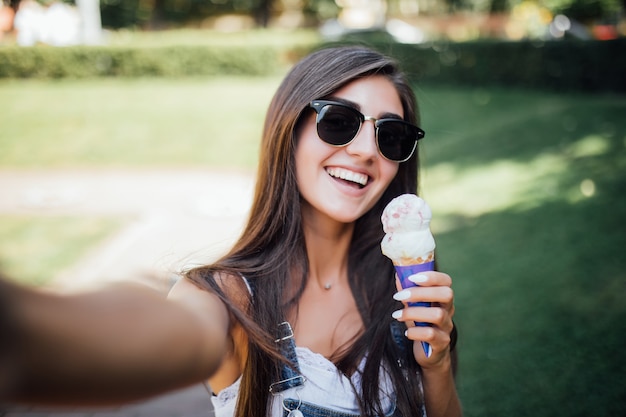 Chica guapa joven hace selfie sonríe con dientes blancos y sostiene el helado con gafas de sol