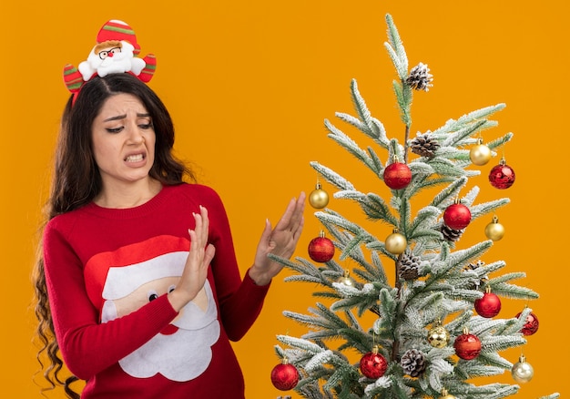 Foto gratuita chica guapa joven irritada con diadema de santa claus y suéter de pie cerca del árbol de navidad decorado mirándolo haciendo gesto de rechazo aislado sobre fondo naranja