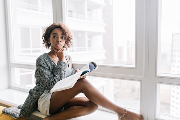 Chica guapa joven con mirada sorprendida sentada en el alféizar de la ventana, gran ventana blanca, leyendo una revista, libro. Lleva chaqueta de punto gris, camiseta, pantalón corto, brazalete de oro en la pierna.