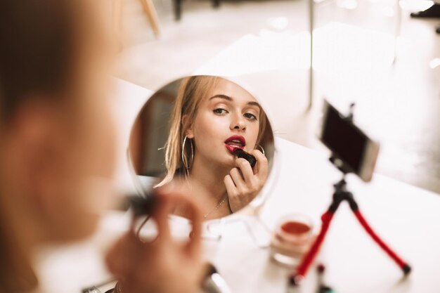 Chica guapa joven mirando en un pequeño espejo en la mesa usando lápiz labial rojo mientras graba un nuevo video para vlog