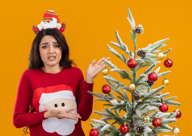 Chica guapa joven molesta con diadema de santa claus y suéter de pie cerca del árbol de navidad decorado apuntando a él mirando a cámara aislada sobre fondo naranja