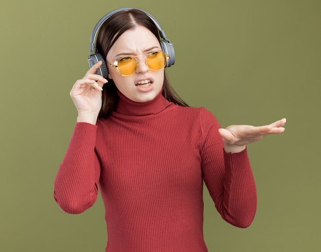 Chica guapa joven molesta con gafas de sol y auriculares mirando al lado tocando los auriculares manteniendo la mano en el aire aislado en la pared verde oliva
