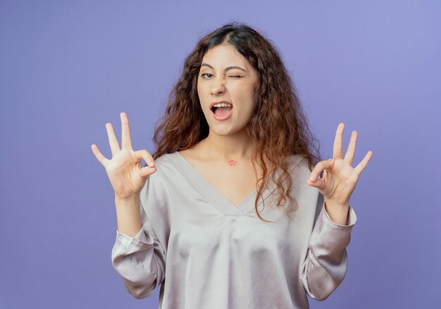 Chica guapa joven parpadeó mostrando gesto okey aislado sobre fondo azul.