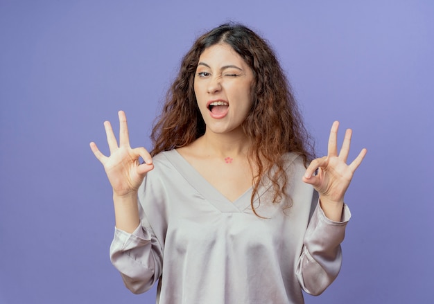 Chica guapa joven parpadeó mostrando gesto okey aislado sobre fondo azul.