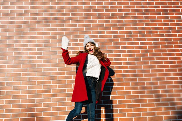 Chica guapa joven con pelo largo en abrigo rojo, sombrero, guantes divirtiéndose en la pared exterior.