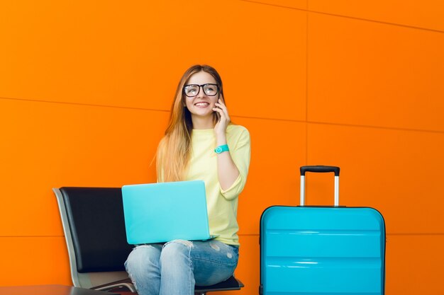 Chica guapa joven con pelo largo está sentada en una silla sobre fondo naranja. Viste suéter amarillo, jeans y lentes. Ella tiene una computadora portátil azul en las rodillas y una maleta. Hablando por teléfono.