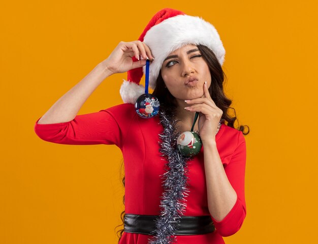 Chica guapa joven pensativa con gorro de Papá Noel y guirnalda de oropel alrededor del cuello sosteniendo adornos navideños mirando al lado tocando la barbilla con un ojo cerrado aislado en la pared naranja