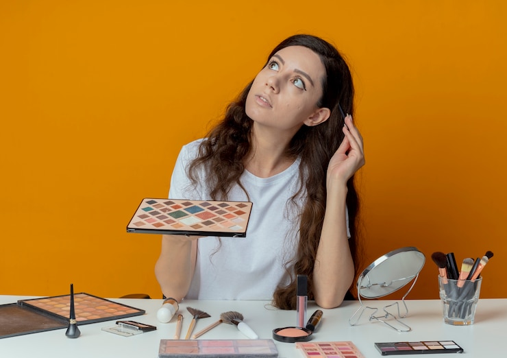 Foto gratuito chica guapa joven pensativa sentada en la mesa de maquillaje con herramientas de maquillaje mirando hacia arriba sosteniendo la paleta de sombra de ojos y tocando la cabeza con un pincel de sombra de ojos aislado sobre fondo naranja